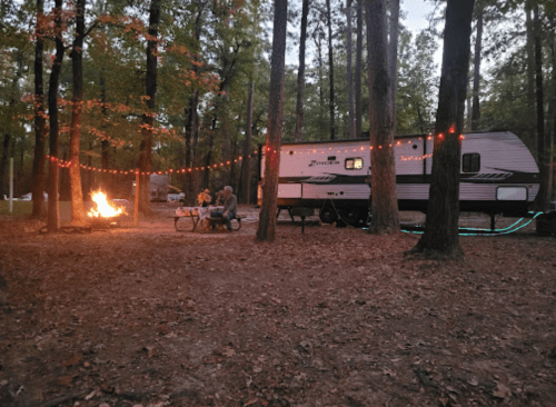 A campsite with a camper, a campfire, and string lights among trees, creating a cozy outdoor atmosphere.