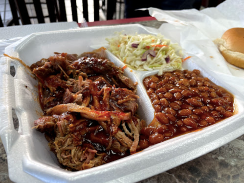 A takeout container with pulled pork in barbecue sauce, baked beans, coleslaw, and a bun on the side.