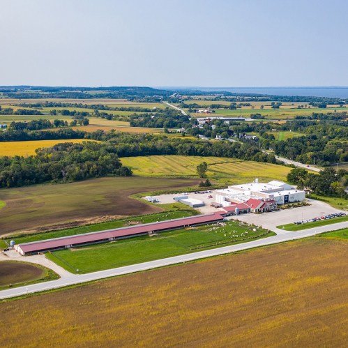 Aerial view of a rural landscape featuring fields, a road, and a building complex surrounded by greenery.