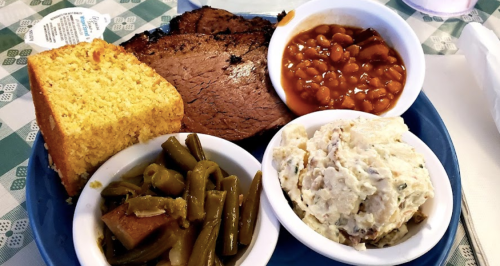 A plate of barbecue brisket, cornbread, baked beans, green beans, and potato salad on a checkered tablecloth.