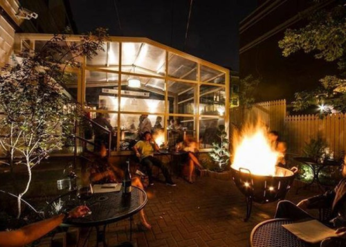 Cozy outdoor seating area at night, featuring a fire pit and a glass-enclosed space with people enjoying the ambiance.