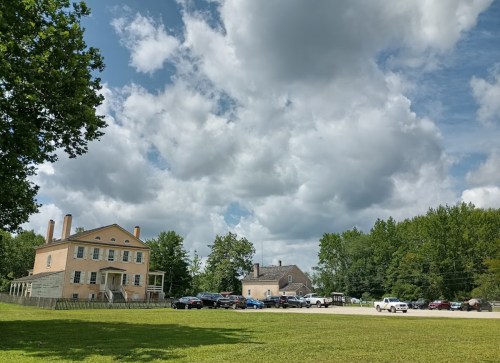 A historic building with a grassy area in front, surrounded by trees and parked cars under a cloudy sky.