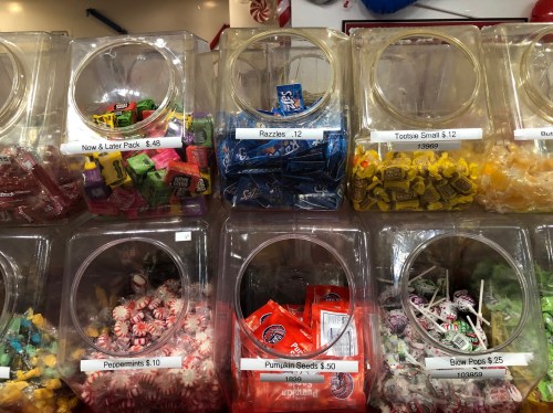 A display of various candies in clear containers, labeled with prices, including Razzles, Tootsie Rolls, and peppermint candies.