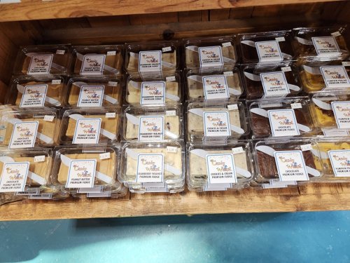 A display of assorted packaged desserts in clear containers, labeled with names and prices, arranged neatly on a wooden shelf.