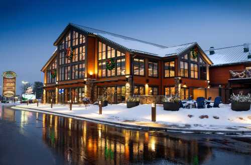 A cozy lodge with large windows, decorated for the holidays, surrounded by snow and reflecting lights on a wet road.