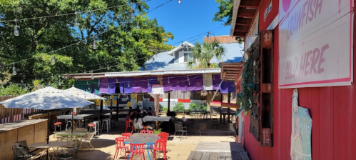 Outdoor dining area with colorful decorations, tables, and string lights, surrounded by trees and a rustic building.