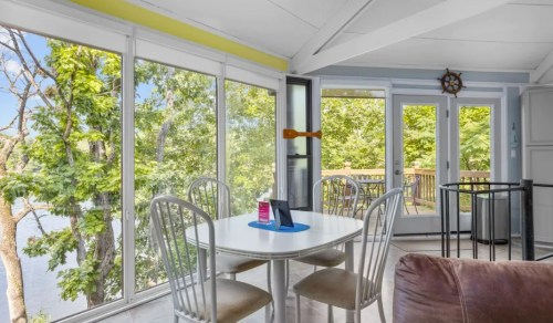 Bright sunroom with a round table, chairs, and large windows overlooking a scenic view of trees and water.