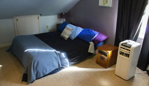 Cozy bedroom with a bed covered in dark bedding, colorful pillows, and a portable air conditioner by the window.
