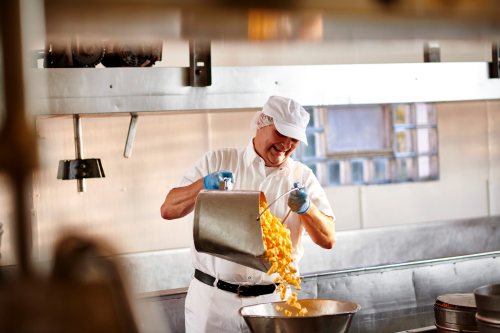 A chef in a white uniform pours pasta from a pot into a large bowl in a kitchen setting.