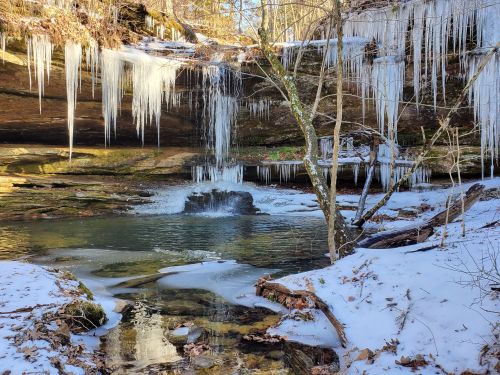 Icicles hang from a rocky ledge above a serene, partially frozen stream surrounded by snow-covered ground and trees.