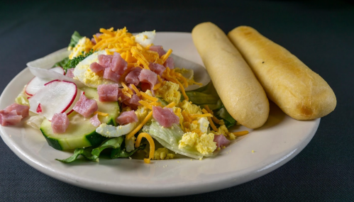 A plate with a salad topped with ham, cheese, and vegetables, accompanied by two breadsticks.