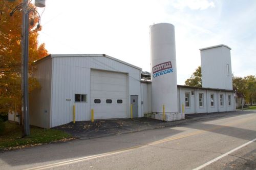 A cheese factory with a tall silo, located on a quiet street, surrounded by trees with autumn foliage.