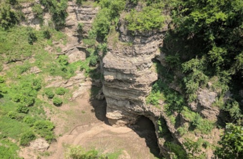 A rocky cliff surrounded by lush greenery, with a dry riverbed visible at the bottom of a deep gorge.