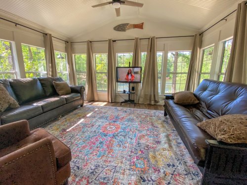 Cozy sunroom with two sofas, a colorful rug, and a TV, surrounded by large windows letting in natural light.