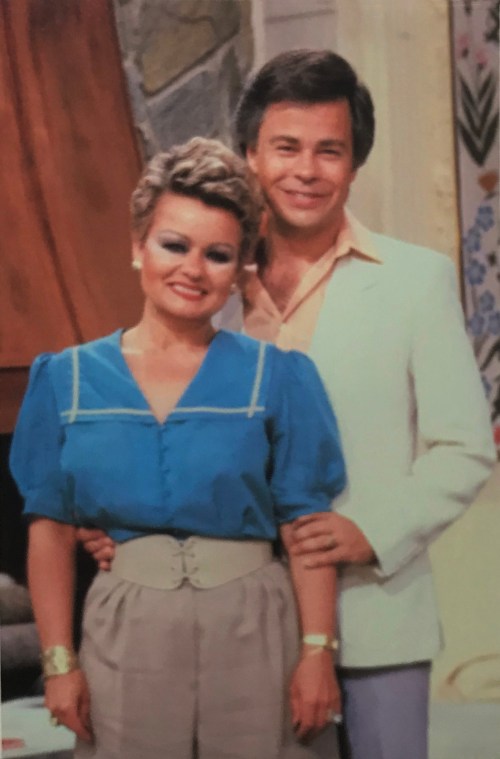 A smiling couple poses together, with the woman in a blue blouse and the man in a white jacket, against a cozy backdrop.
