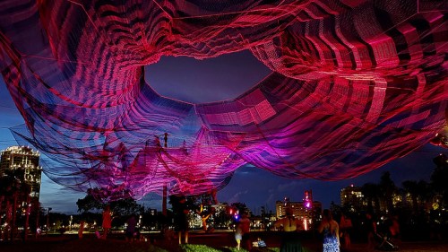 A vibrant, colorful net installation hangs above a park at dusk, illuminated by pink and purple lights.