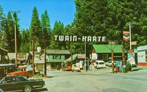 A vintage view of Twain-Harte, featuring a sign, shops, and classic cars surrounded by tall trees.