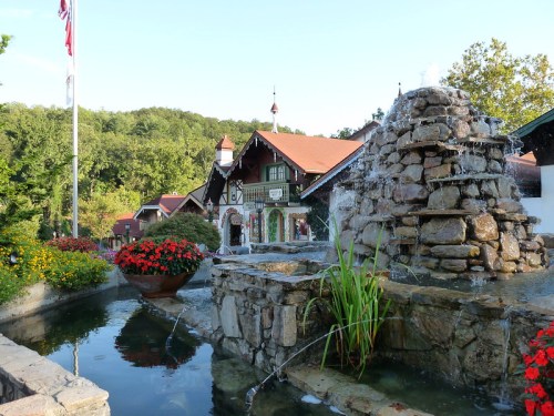 A stone fountain with water flowing, surrounded by colorful flowers, in front of a charming building and green hills.