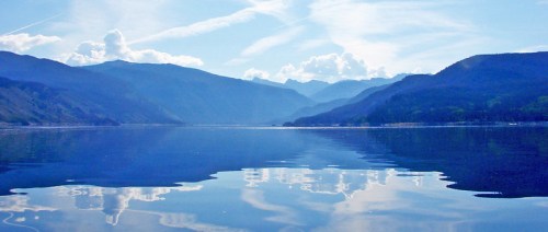 A serene lake reflecting mountains and a blue sky with fluffy clouds, creating a tranquil landscape.