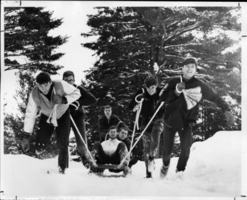 A group of six people pulls a sled through the snow, surrounded by trees on a winter day.