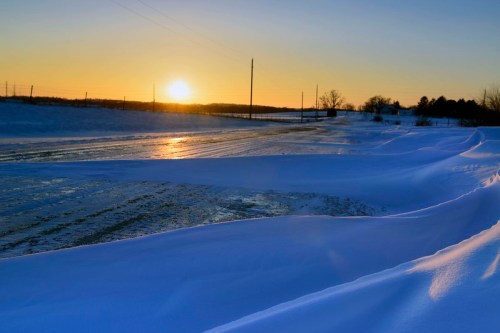 A snowy landscape at sunset, with a winding road and soft snowdrifts under a clear sky.