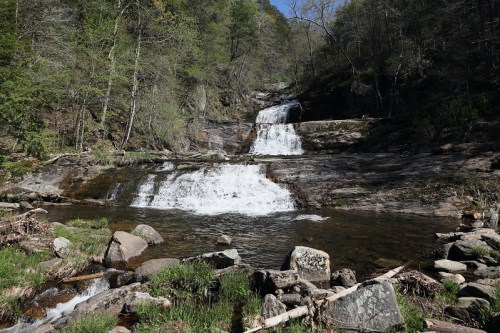 A serene waterfall cascades over rocky terrain, surrounded by lush greenery and trees under a clear blue sky.