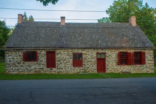 A historic stone house with a shingled roof, red doors, and windows, set against a green landscape.