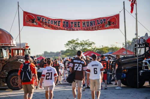 A crowd of fans in Browns jerseys walking under a banner that reads "Welcome to the Muni Lot" at a football event.