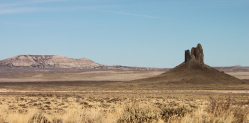 A rocky formation rises from a vast, arid landscape under a clear blue sky, with distant mountains in the background.