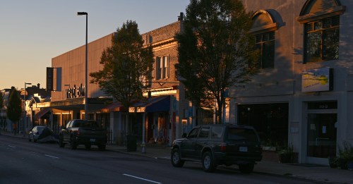 Street view at sunset, featuring shops and parked cars along a quiet road lined with trees.