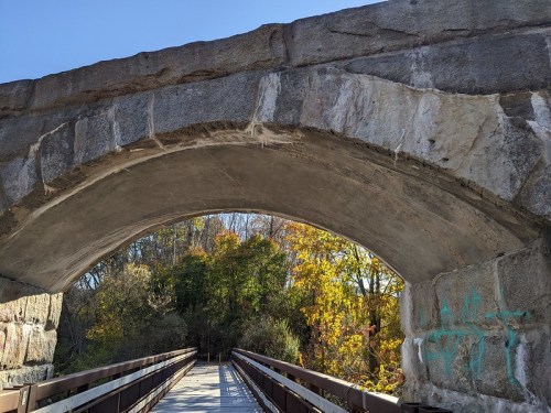 View of a stone arch bridge with a wooden walkway, surrounded by trees displaying autumn colors.