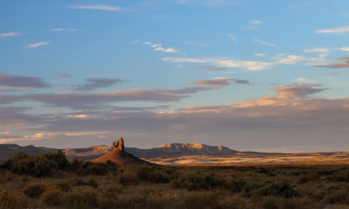 A scenic landscape featuring a rocky formation under a blue sky with scattered clouds and distant mountains.