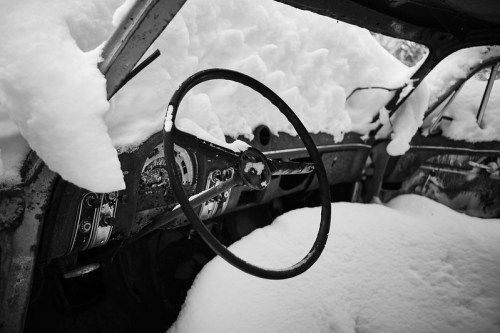 An abandoned car interior covered in snow, featuring a steering wheel and dashboard in a monochrome setting.