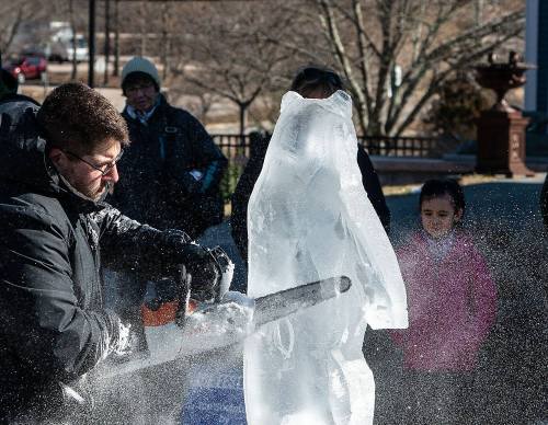 A man carves an ice sculpture of a bear with a chainsaw, while a girl watches nearby in a winter setting.