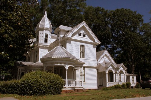 A large, ornate white Victorian house with a turret and intricate architectural details, surrounded by greenery.