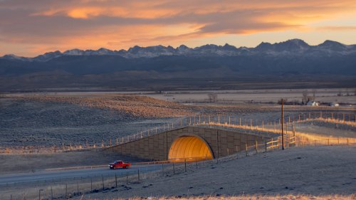 A red car drives past a golden tunnel under a sunset, with mountains in the background and open fields surrounding.