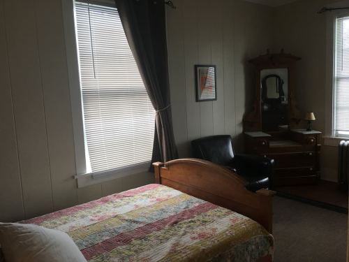 A cozy bedroom featuring a bed with a colorful quilt, a chair, and a dresser with a mirror, illuminated by natural light.