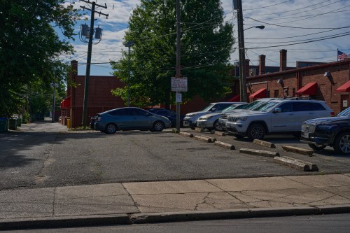 A parking lot with several cars, surrounded by trees and buildings, under a partly cloudy sky.
