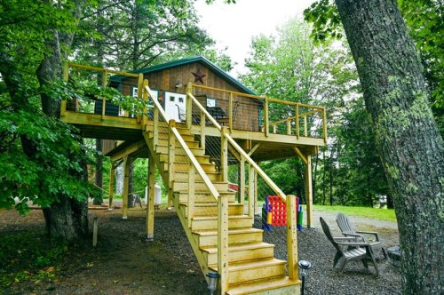 A wooden cabin on stilts surrounded by trees, featuring a staircase and outdoor seating area.