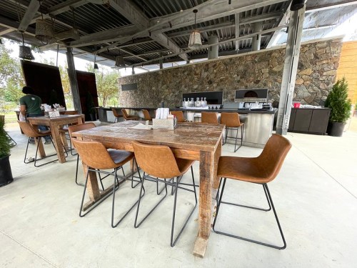 Outdoor dining area with wooden tables and brown chairs, featuring a stone wall and a bar in the background.