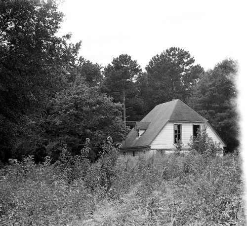 An abandoned house with a triangular roof, surrounded by overgrown grass and trees in a black-and-white setting.