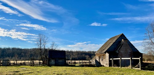 A rustic, weathered house and a small shed under a blue sky with scattered clouds, surrounded by a grassy field.