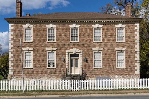 A historic brick house with white trim and a picket fence, featuring multiple windows and a central entrance.