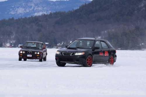 Two cars racing on a snowy landscape, with mountains in the background. One car is black with red wheels, the other is gray.