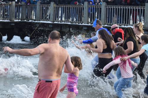 A group of people, including children, splash into a snowy river, with onlookers watching from a nearby pier.