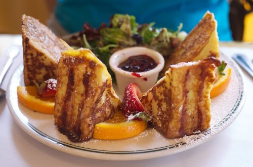 A plate of French toast slices with strawberries, orange slices, and a side of mixed greens and berry sauce.
