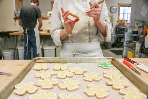 A baker decorates cookies on a tray, with a blurred figure working in the background.