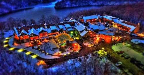 Aerial view of a cozy lodge surrounded by snow, illuminated by warm lights, near a river and wooded area.