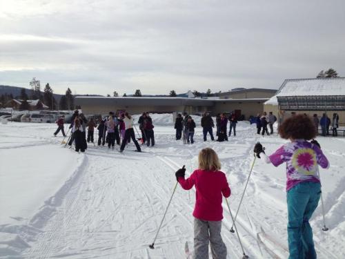 A group of people, including children, skiing on a snowy trail with a building in the background.