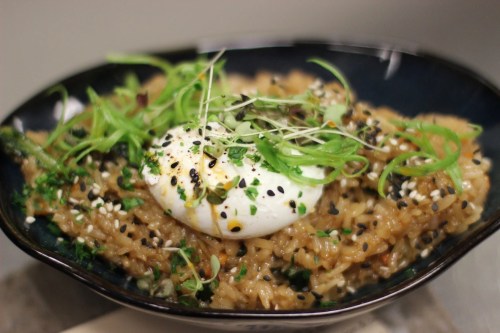 A bowl of risotto topped with a poached egg, green onions, and sesame seeds, garnished with herbs.